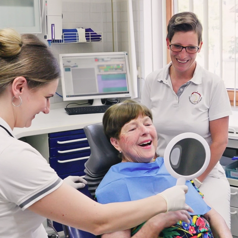 Aufnahme einer Patientin, die während sie im Beisein von Zahnärztin Sarah Knefel und Assistentin in den Handspiegel lächelt.